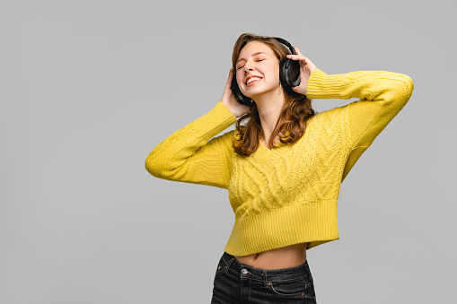 Woman is using wireless modern headphones and listening music. Portrait of nice person who posing at the studio with grey background. Concept with real people.