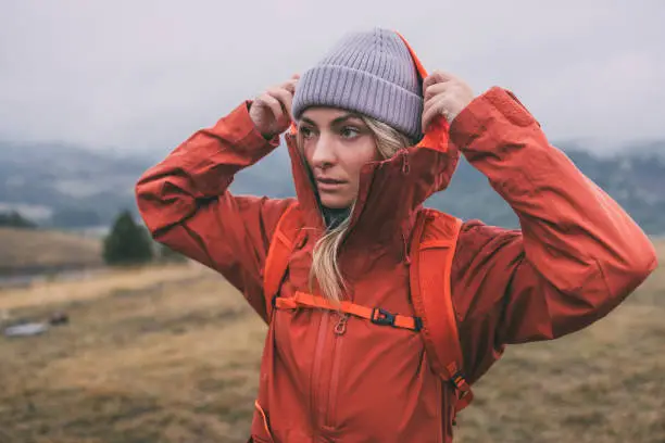 Photo of Portrait of a Beautiful Blonde Female Hiker in a Red Jacket and a Gray Hat, Exploring the Mountains on a Cloudy Day