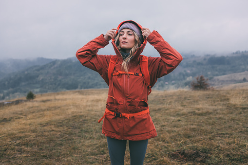 A young female explorer wearing a red jacked and a backpack, wandering in the meadows of a mountain on a cloudy day