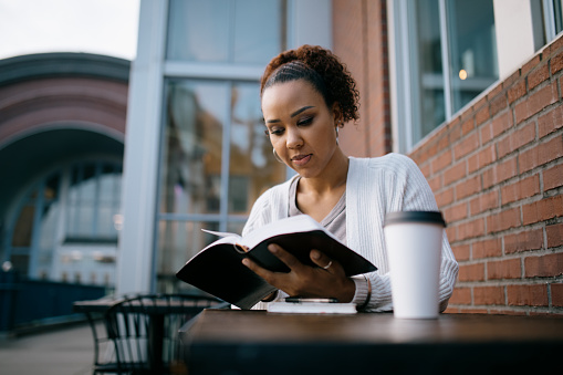 A mixed race women reads a large Bible / textbook while seated at table outdoors.  Could also be a university student on campus.