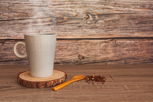 On a table is a mug with a hot drink placed on a coaster made from the slice of a trunk. There is a small wooden spoon and a bunch of herbs. In the background are wooden planks.