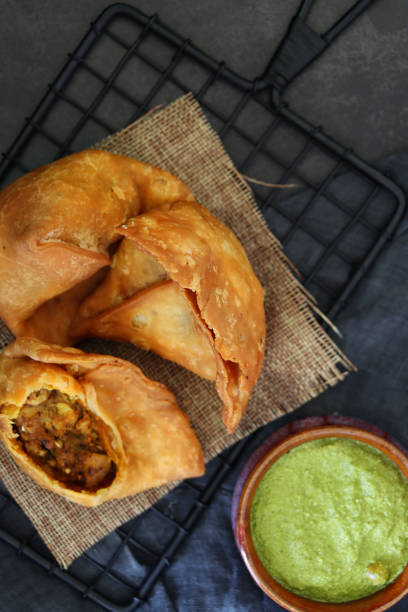 istock Image of cooling rack with hessian full of fried samosas, samosa with missing bite stuffed with spiced potato, peas and meat, bowl of mint coriander dip, grey mottled background, elevated view 1349831642