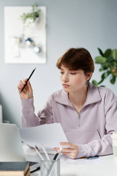 istock Thoughtful young Caucasian marketing specialist in sweatshirt using laptop and analyzing papers while preparing new campaign 1349835197