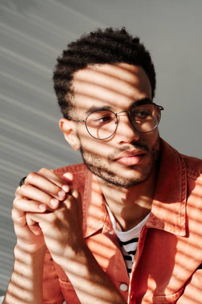 istock Pensive young black man with stubble wearing round glasses looking away, light through blinds effect 1349836949