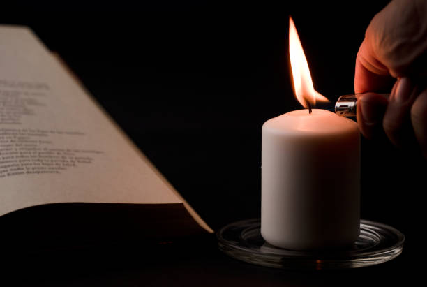 istock candles with flame, man's hand with lighter, and reading book on desk, black background. (focus on candle). 1349837008