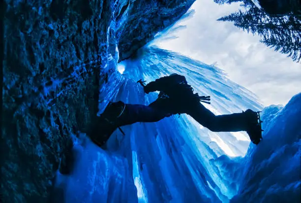 Ice Climber from Below with Dramatic Ice - Epic view with blue icicles and climber silhouette. Ice climbing in mountains wide angle view. Extreme outdoor adventure sports.