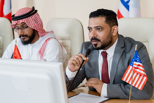 A photo of confident Arab businessman discussing in a meeting with middle eastern business partner. Emirati man wearing traditional clothes. Professional is gesturing while sitting with colleague at conference table, at brightly lit office.
