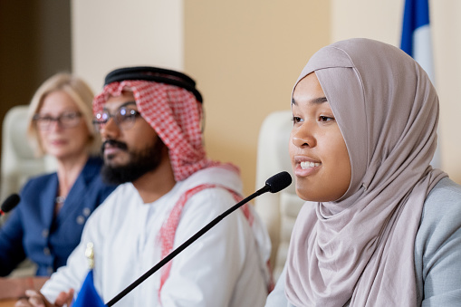 Attractive young muslim woman in hijab speaking into microphone while participating in conference discussion