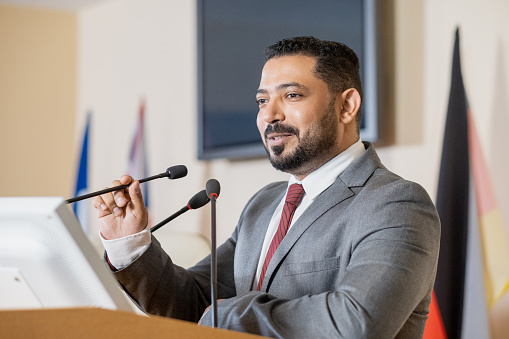 Content confident Middle Eastern politician in gray suit standing at tribune podium and speaking into microphone while presenting report at conference