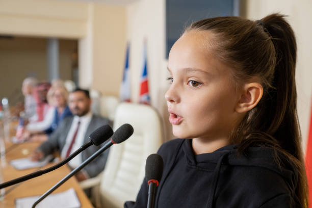 Little Activist Addressing Conference Little activist with ponytail speaking into microphone while addressing conference in front of congress audience activist speech stock pictures, royalty-free photos & images