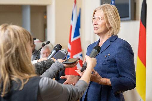 Confident blond-haired congresswoman gesturing hands while answering questions of journalists with microphones