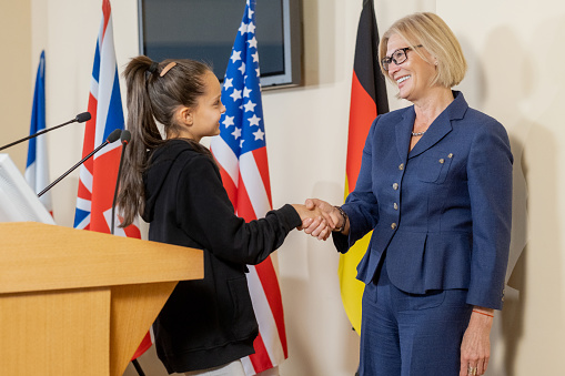 Smiling mature congresswoman in formal suit shaking hand of little activist after her presentation at political meeting