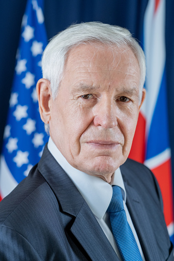 Portrait of serious senior Caucasian congressman in formal suit standing against national flags