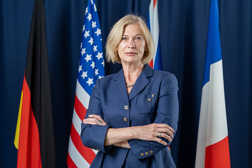 Portrait of serious confident mature female politician in dark blue jacket standing with crossed arms against national flags
