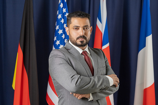 Portrait of serious confident middle eastern political candidate in suit standing with crossed arms against international flags