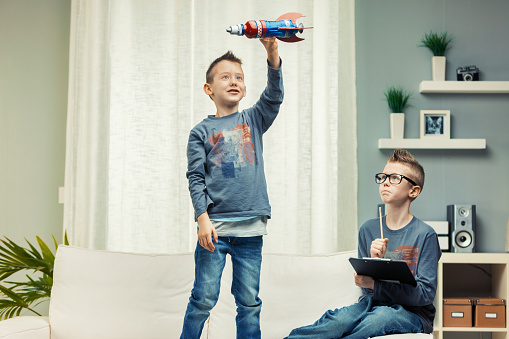 Two cute young boys playing together at home, one with tablet and one flying toy rocket in air