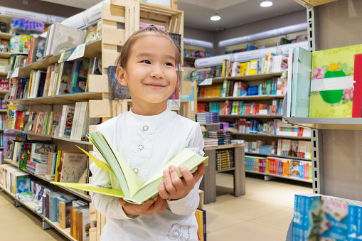 child girl stands in the middle of bookshelves holding a book in her hands and looks, smiling to the side. Bottom view.
