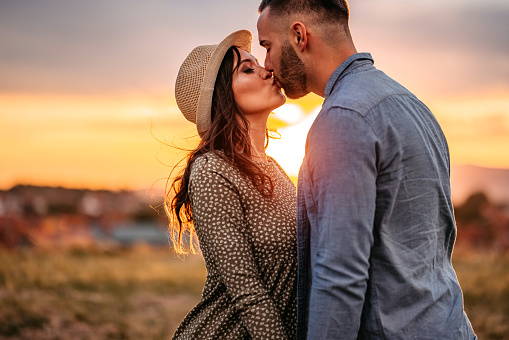 Young happy cute pregnant couple kissing on meadow at sunset.