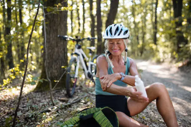 A happy senior woman biker sitting and applying sunscreen on her arm outdoors in forest.