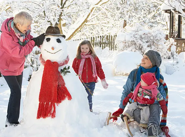 Young Girl With Grandmother And Mother Building Snowman In Garden Having Fun Laughing