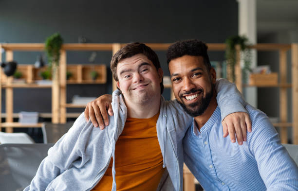 Young man with Down syndrome and his tutor with arms around looking at camera indoors at school Happy young man with Down syndrome and his tutor with arms around looking at camera indoors at school down syndrome photos stock pictures, royalty-free photos & images