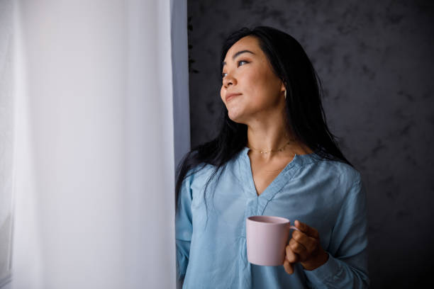 istock Happy young woman looking through the window and contemplating while having morning coffee 1349909123