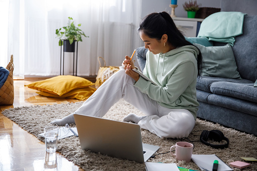 Wide shot of diligent young woman sitting on the living room floor, studying for university and writing homework in her notebook.