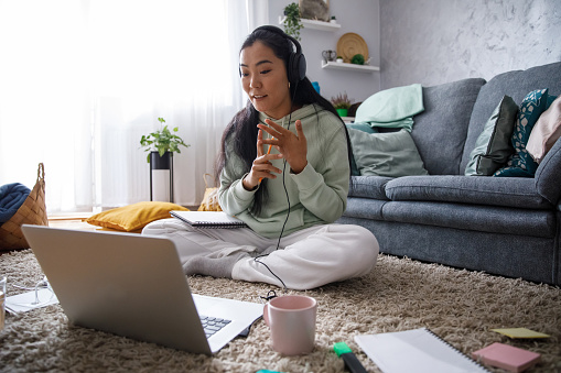 Wide shot of intelligent female university student sitting on the living room floor and counting on fingers a subject matter she is discussing with her classmates and professor during online class via video conference call on laptop.