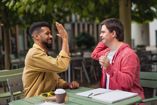 Young man with Down syndrome with mentoring friend sitting outdoors in cafe celebrating success. stock photo