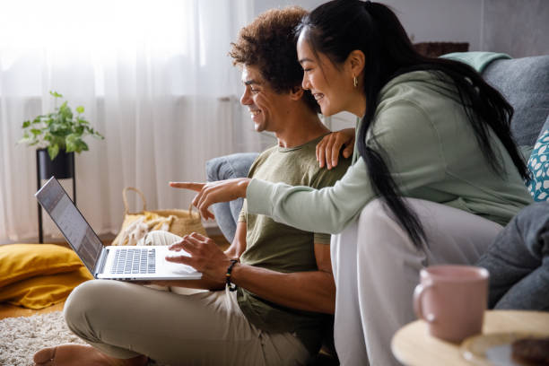 Young woman pointing at something on laptop screen while looking at online banking with her boyfriend Copy space shot of young man sitting on the floor and using laptop, organizing finances and online banking. His girlfriend is sitting on the sofa, behind him, and pointing at something on the screen. central asian ethnicity photos stock pictures, royalty-free photos & images
