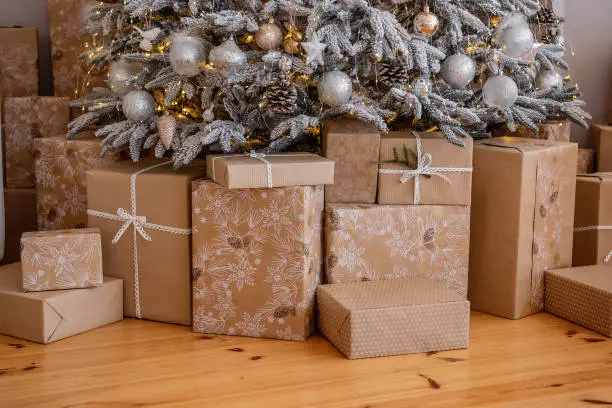 Photo of On wooden floor under snowy artificial Christmas tree, boxes in craft paper with vintage ribbon