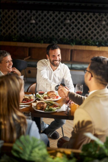 Successful deal on a business lunch! Happy businessmen shaking hands while having a lunch with their females colleagues in a restaurant. business lunch stock pictures, royalty-free photos & images