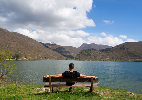 A person sitting from behind contemplates the lake on a beautiful day with some clouds in the sky