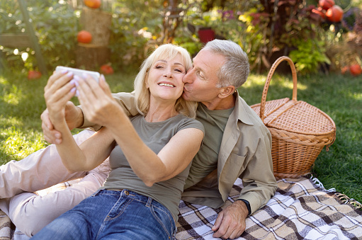 Happy senior couple taking selfie on smartphone, having picnic and resting in garden outside, enjoying autumn nature, copy space. Elderly spouses capturing happy moments