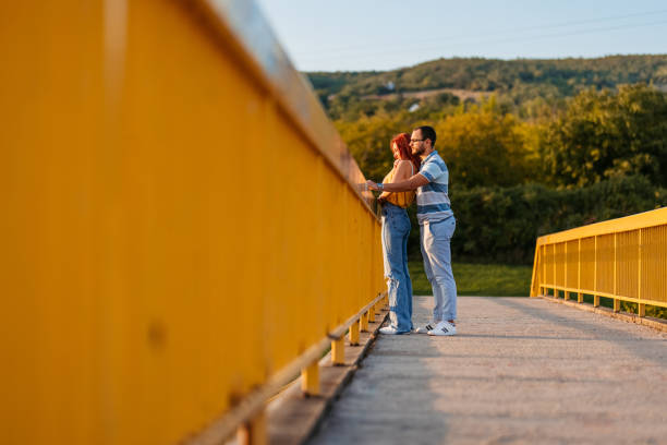 Young heterosexual loving couple leaning on fence and enjoying at view from bridge.