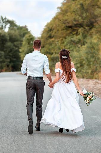 The newlyweds hold hands and run along the road. View from the back. Wedding and Day concept.