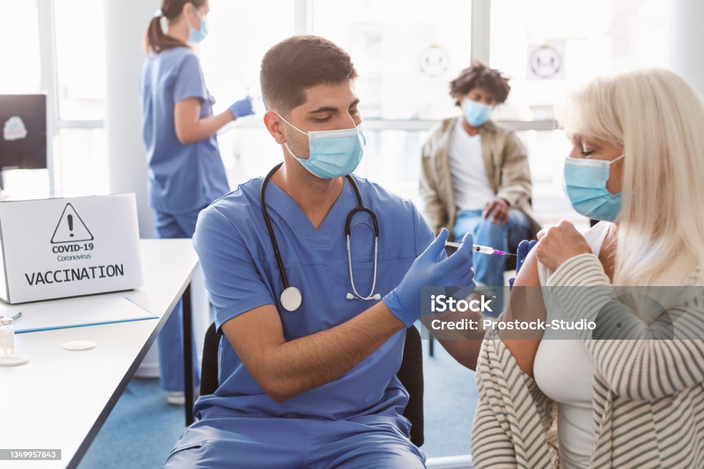 Mature Lady Receiving Vaccine Injection In Arm In Health Center Male doctor holding syringe making covid 19 vaccination injection dose in shoulder of elderly female patient. Flu influenza vaccine clinical trials, corona virus treatment side effect, inoculation Vaccination Stock Photo