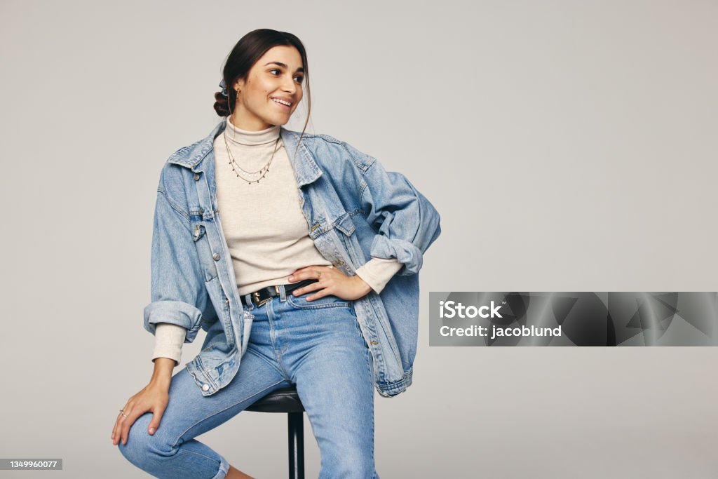 Rocking denim wear Rocking denim wear. Fashionable young woman sitting on a chair against a grey background. Happy young woman looking away with a smile on her face in a modern studio. Jeans Stock Photo