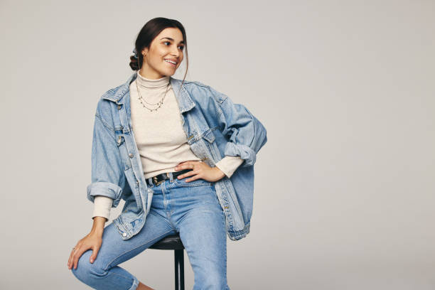 Rocking denim wear Rocking denim wear. Fashionable young woman sitting on a chair against a grey background. Happy young woman looking away with a smile on her face in a modern studio. denim stock pictures, royalty-free photos & images
