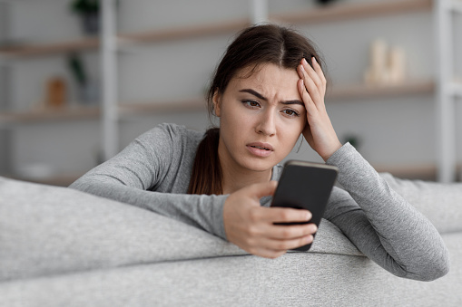 Sad frustrated shocking european young woman suffering from depression and reading message with bad news. Jealous lady sit on sofa hold phone waiting for call, upset or worried at home, empty space