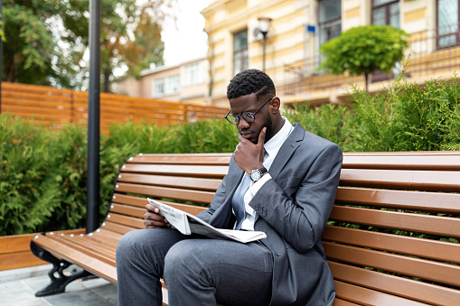 African american businessman in suit reading newspaper while sitting on bench near office center, free space. Thoughtful black office worker reading daily news