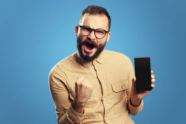 Excited young man with glasses screaming and showing empty screen smartphone while celebrating victory with raised fist, isolated over blue backdrop