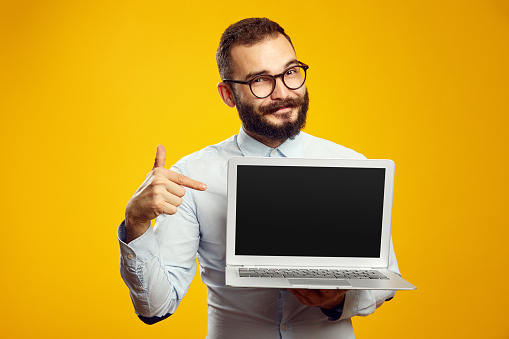 Cheerful hipster man with beard wearing beige shirt and eyeglasses pointing at new laptop in hands on yellow background.