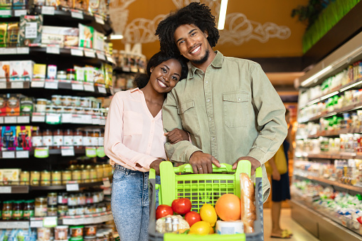 Cheerful Black Couple On Grocery Shopping, Posing With Shop Cart, Buying Food Products Together In Store, Smiling To Camera. African American Family In Modern Supermarket Concept.