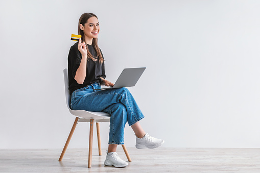 Pretty young woman with credit card and laptop sitting on chair, ordering goods on internet, buying things in web store against white wall, copy space. Online shopping, remote banking concept