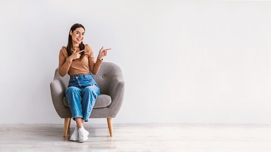 Cool offer. Happy young lady sitting in armchair, pointing aside at free space against white studio wall, banner design. Positive millennial woman offering place for advert or promotional text