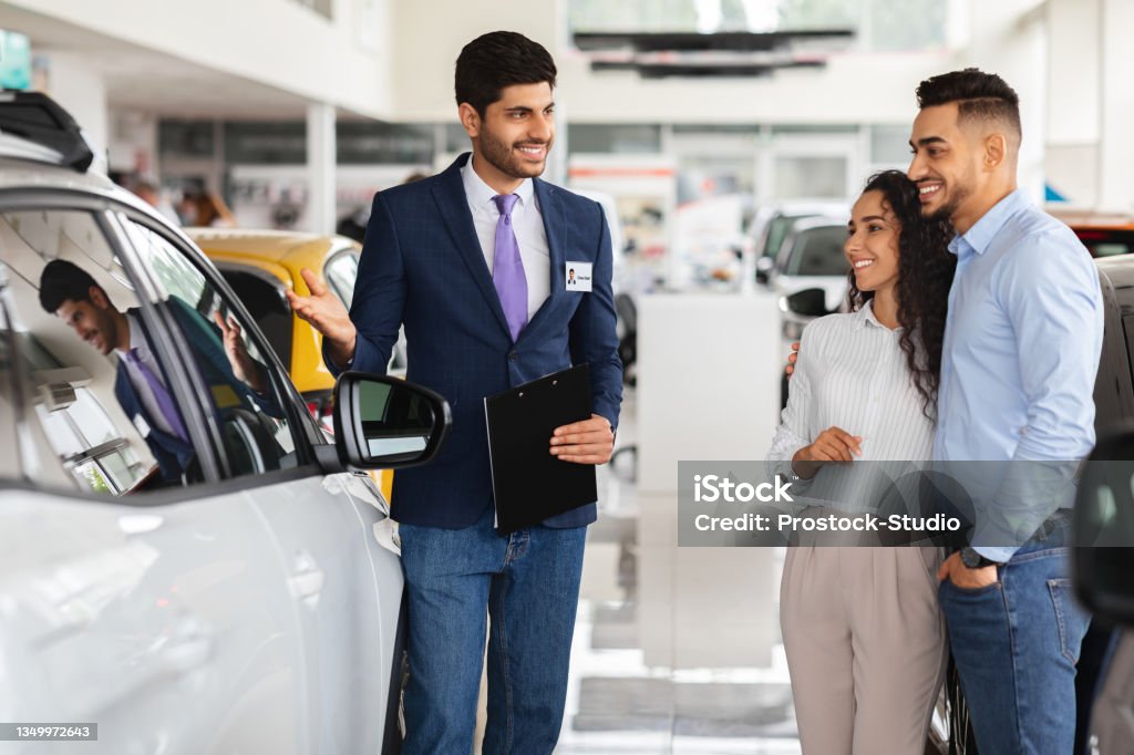 Smiling middle-eastern couple looking at white car Smiling middle-eastern couple looking at brand new white car, listening to friendly sales manager talking about auto specifications, luxury dealership salon interior, side view, copy space Car Dealership Stock Photo