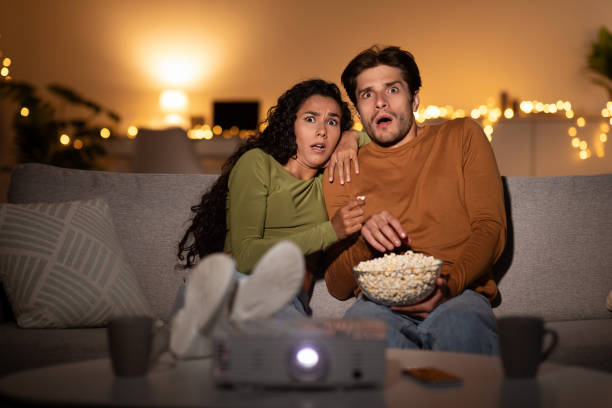 istock Scared Couple Watching Horror Movie In Shock Using Projector Indoors 1349972698