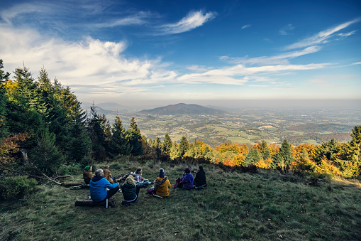 Two families with teenage kids hiking together in autumn forest. They are sitting on a tree trunks, resting on meadow and enjoying the spectacular view over the countryside.\nShow with Canon R5