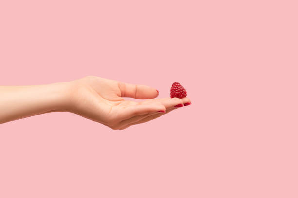 istock Young female hand hold ripe red raspberry on a fingers. Female hand with raspberry isolated on pink background. 1349979776